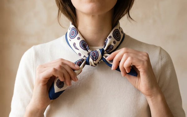 Hands tying a navy and ivory medallion silk square scarf at the neck