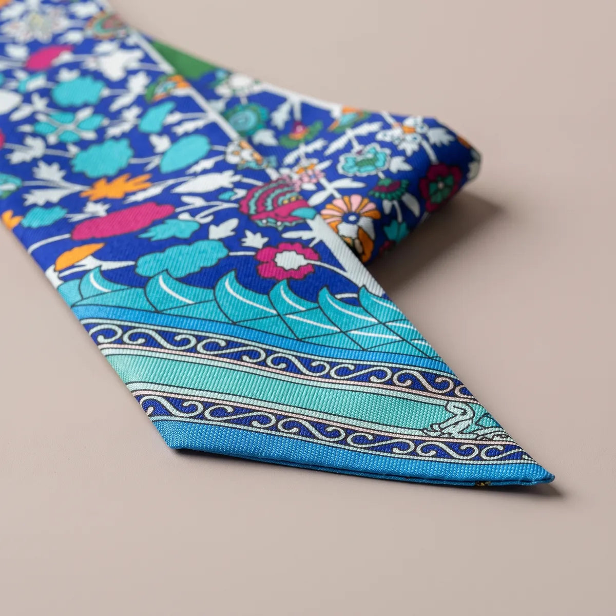 Slanted tip of the blue skinny scarf with turquoise wave bands, a running scroll and tiny red flowers above