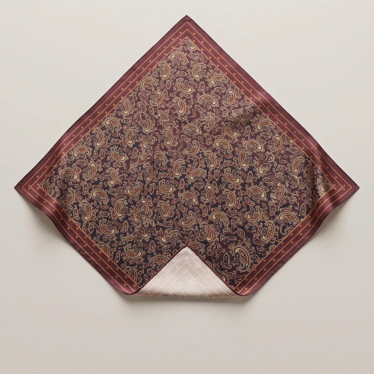 Burgundy paisley silk bandana laid flat, paisleys on navy and wine, striped border, one corner turned to the pale reverse