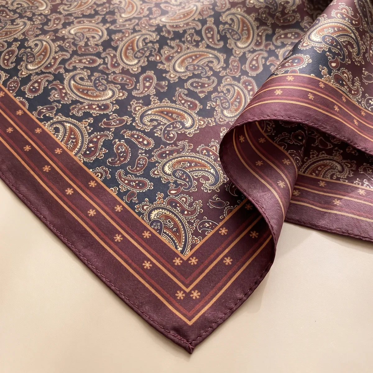 Corner detail of the bandana: rust, tan and cream paisleys on navy beside a burgundy border with small stars