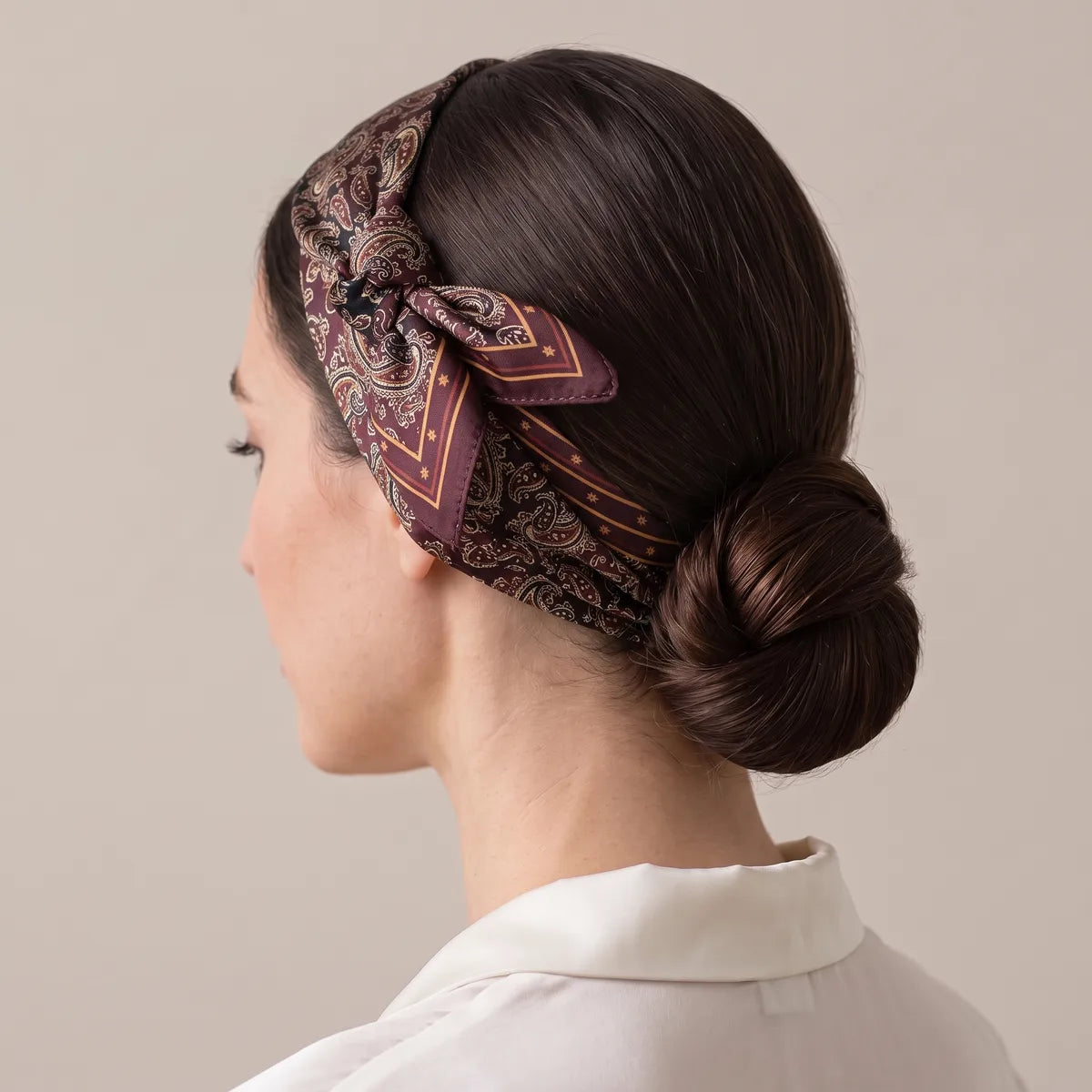 Paisley silk bandana in burgundy folded into a headband, knotted high on the side with two pointed ends, worn with a low bun