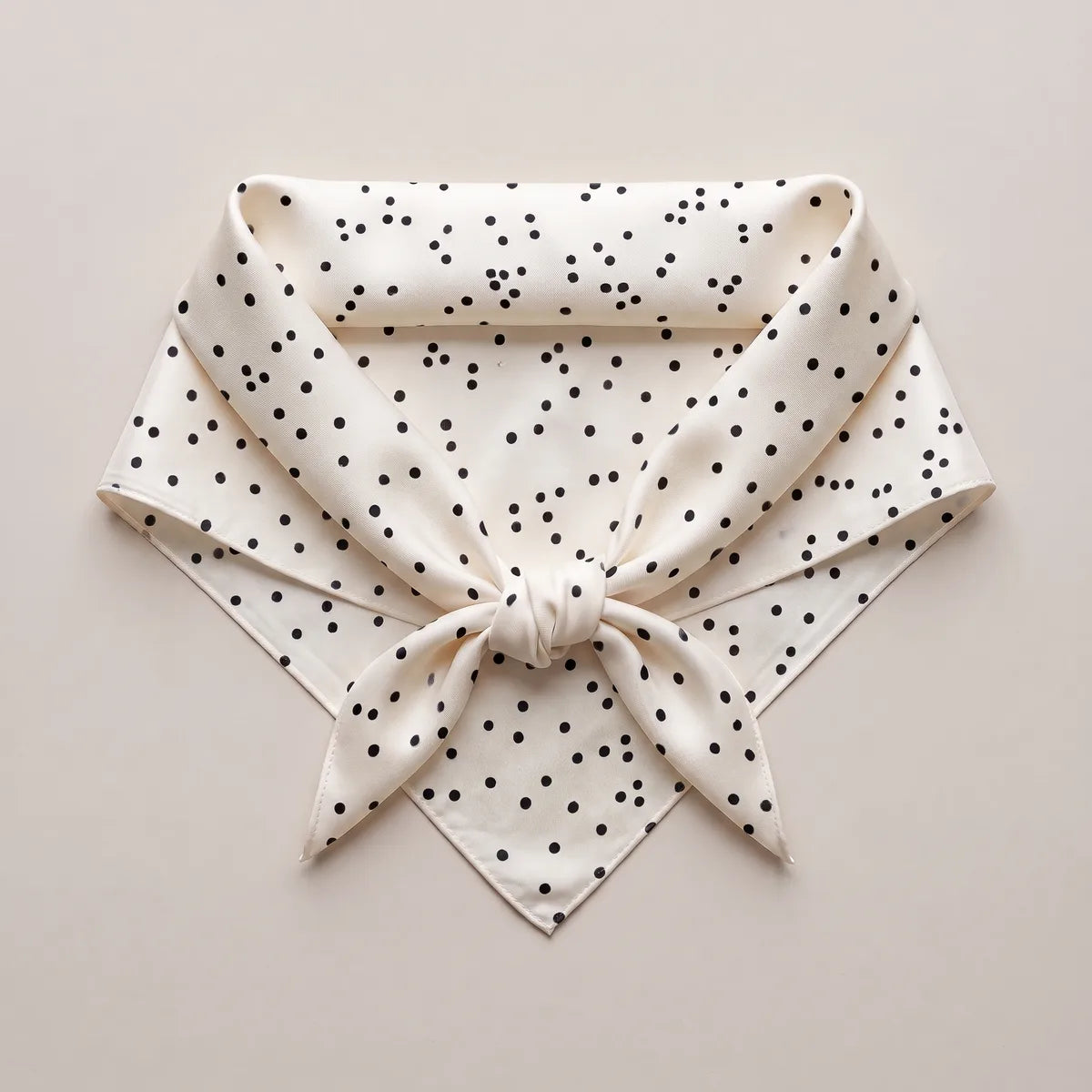 Cream silk square with black dots knotted into a neckerchief shape and laid flat, dotted points spread below