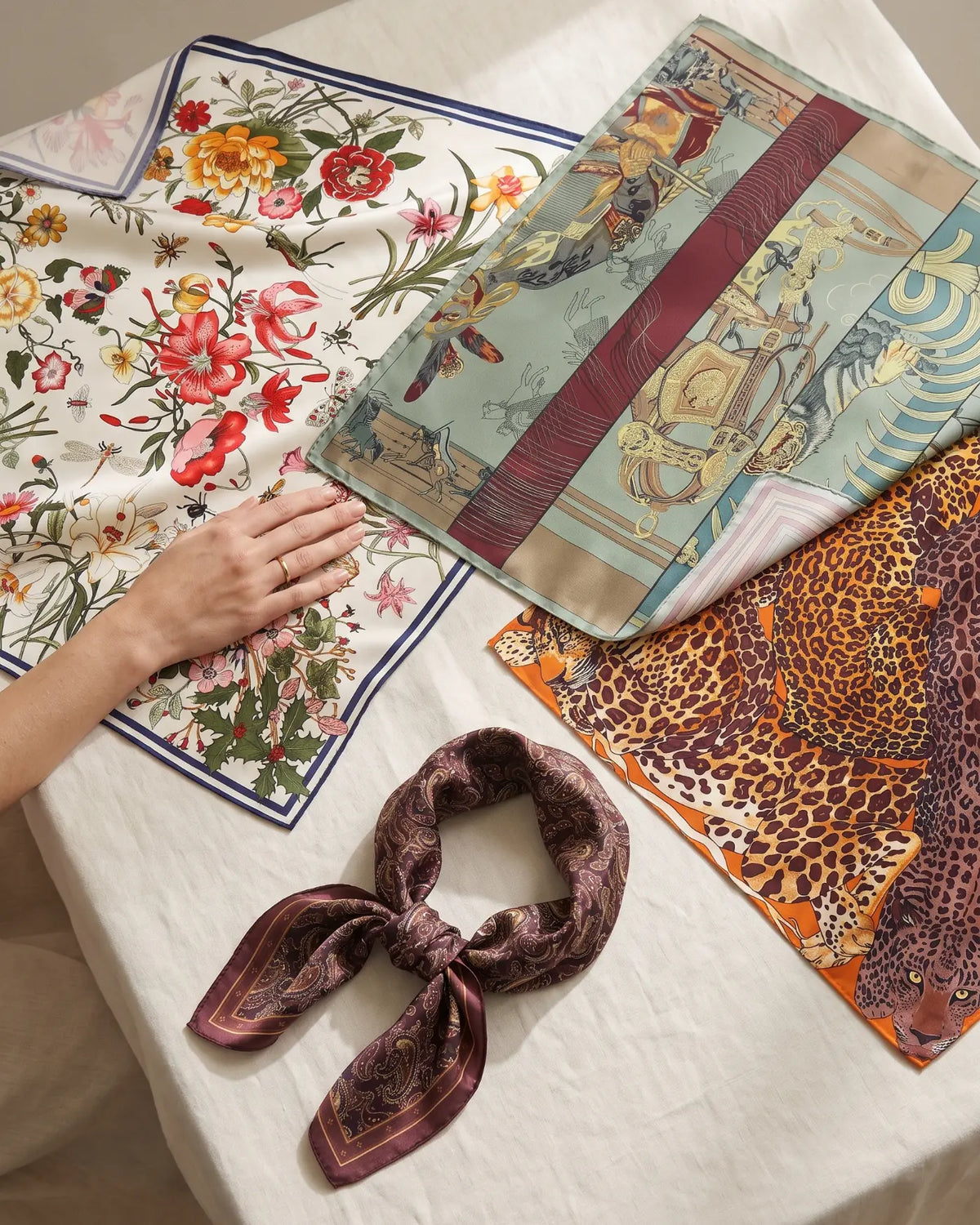 Hand choosing between floral, equestrian and leopard print silk scarves, with a paisley bandana tied in a knot