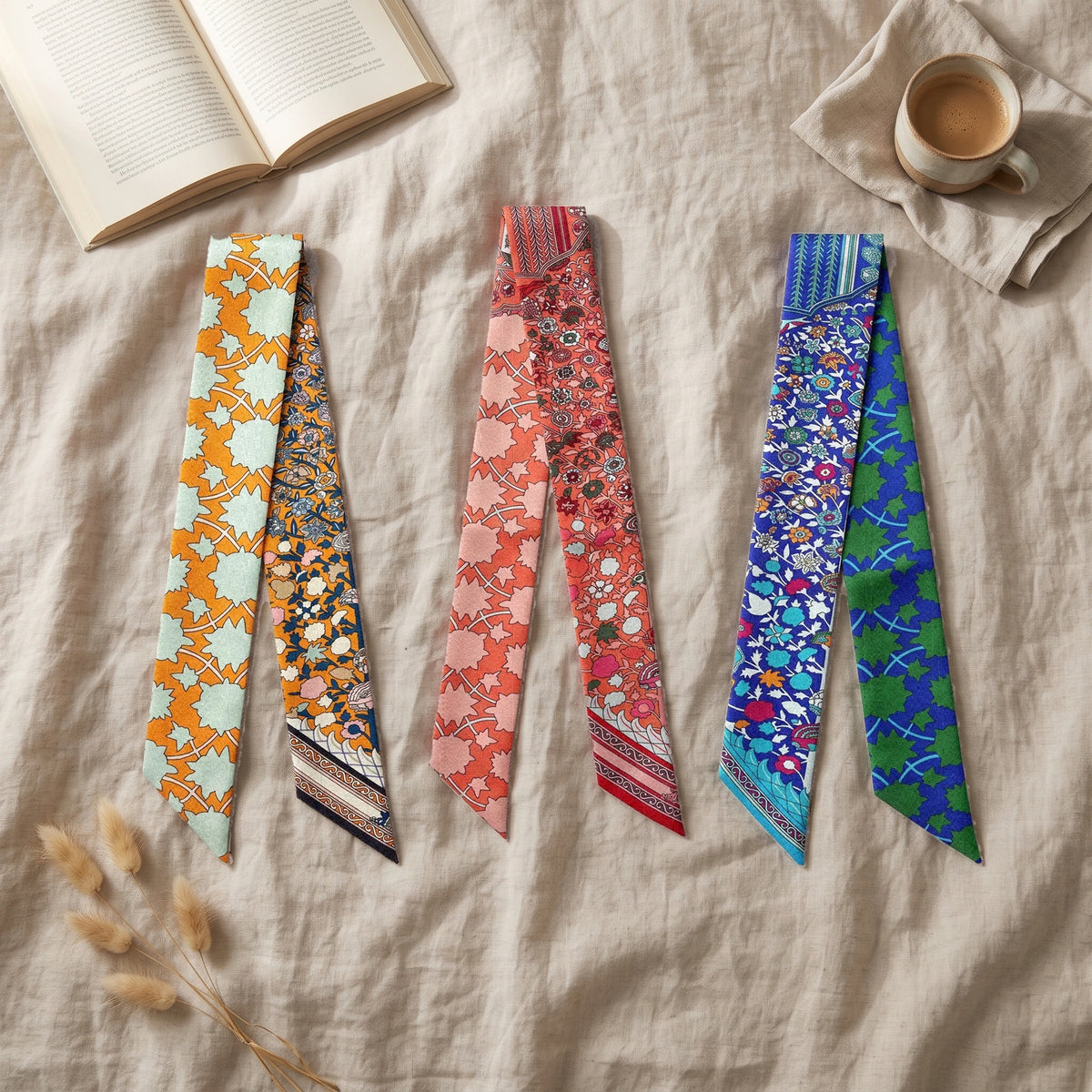 Floral skinny scarves in orange, coral and cobalt spread across a linen sheet with a coffee and a book