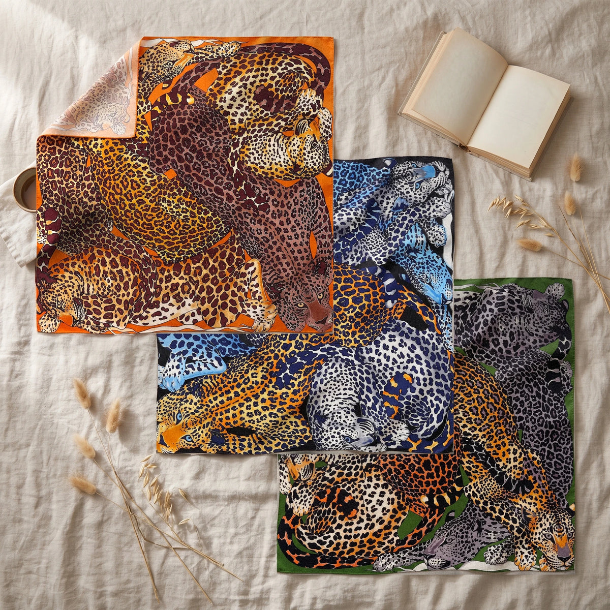Three leopard print shawls fanned out on a linen bedspread with an open book, a mug and sprigs of dried grass