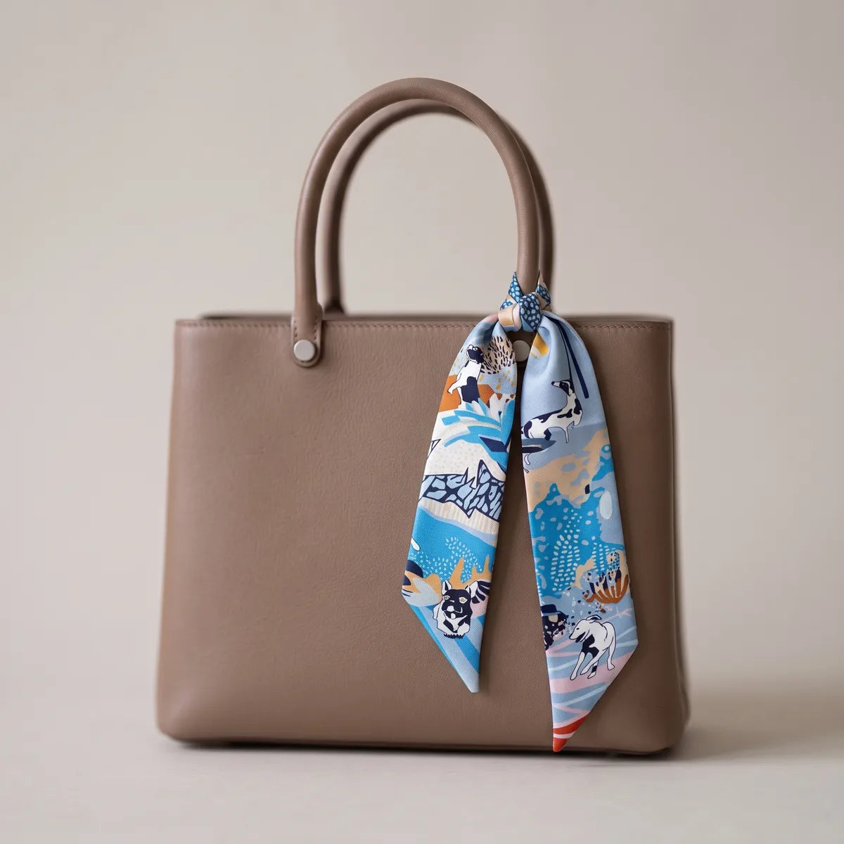 Light blue dog print skinny silk scarf hanging from a taupe handbag handle, red tip at the end
