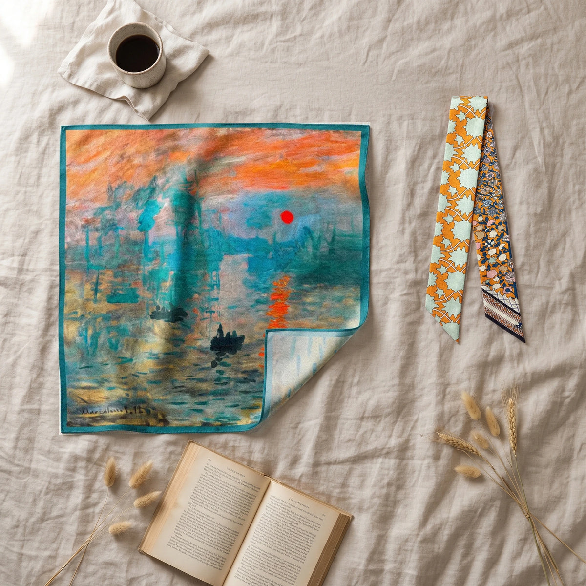 Teal sunrise bandana and orange floral skinny scarf laid on linen next to a coffee cup and a book
