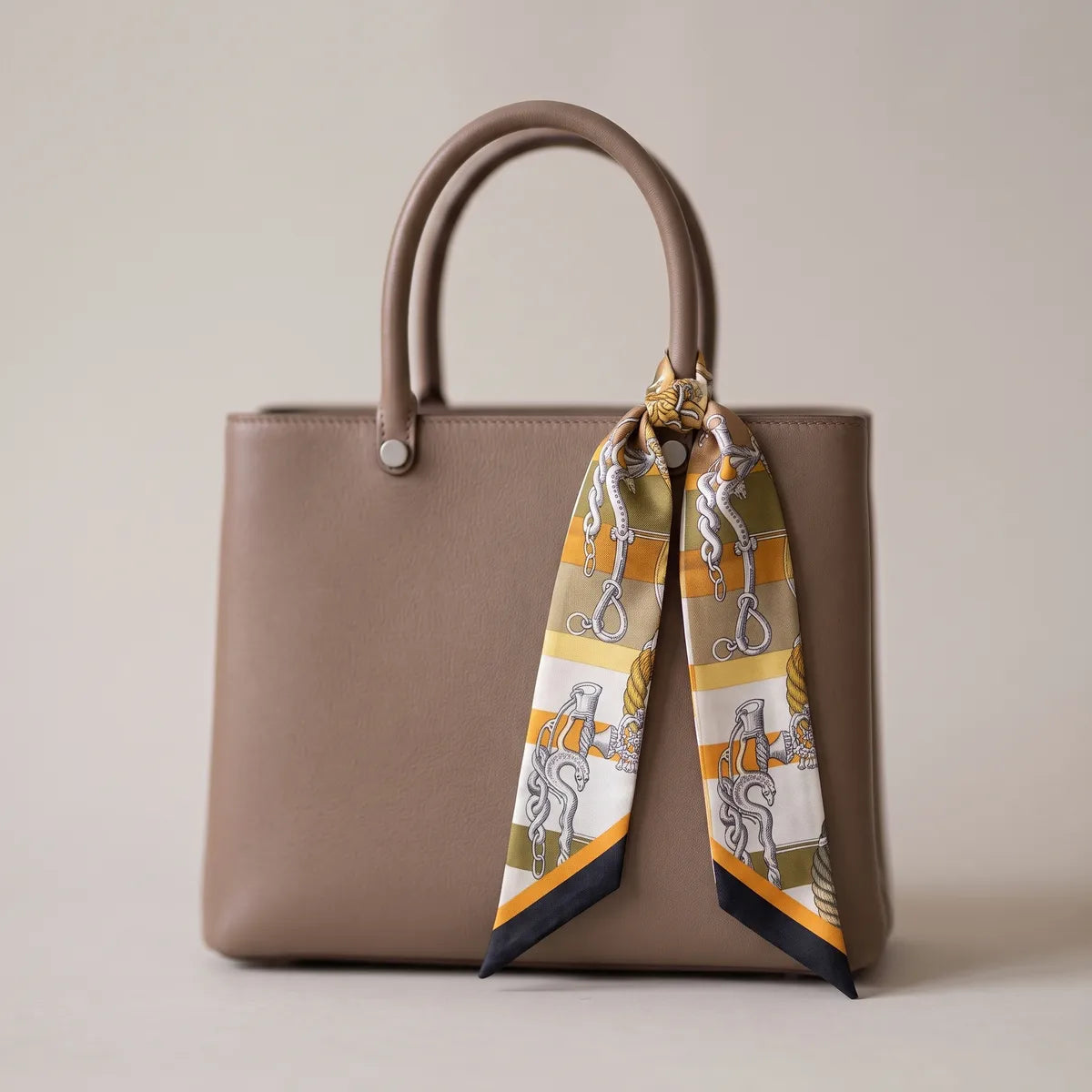 Mustard chain print skinny silk scarf tied to the handle of a taupe leather bag