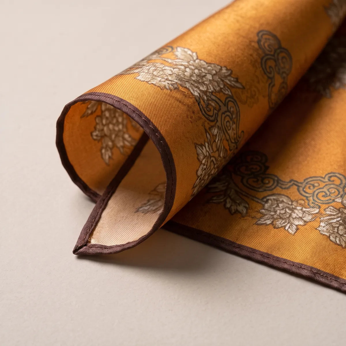 Rolled corner of the mustard floral scarf revealing the pale sand-colored reverse and the brown edge