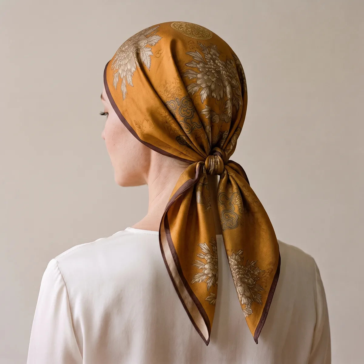 Mustard silk scarf seen from behind, covering the whole head, knot at the nape and brown-edged tails