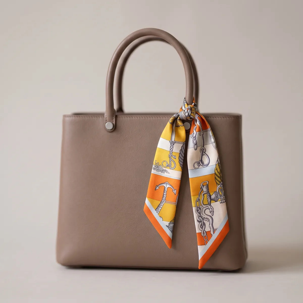 Orange chain print skinny silk scarf knotted on the handle of a taupe handbag