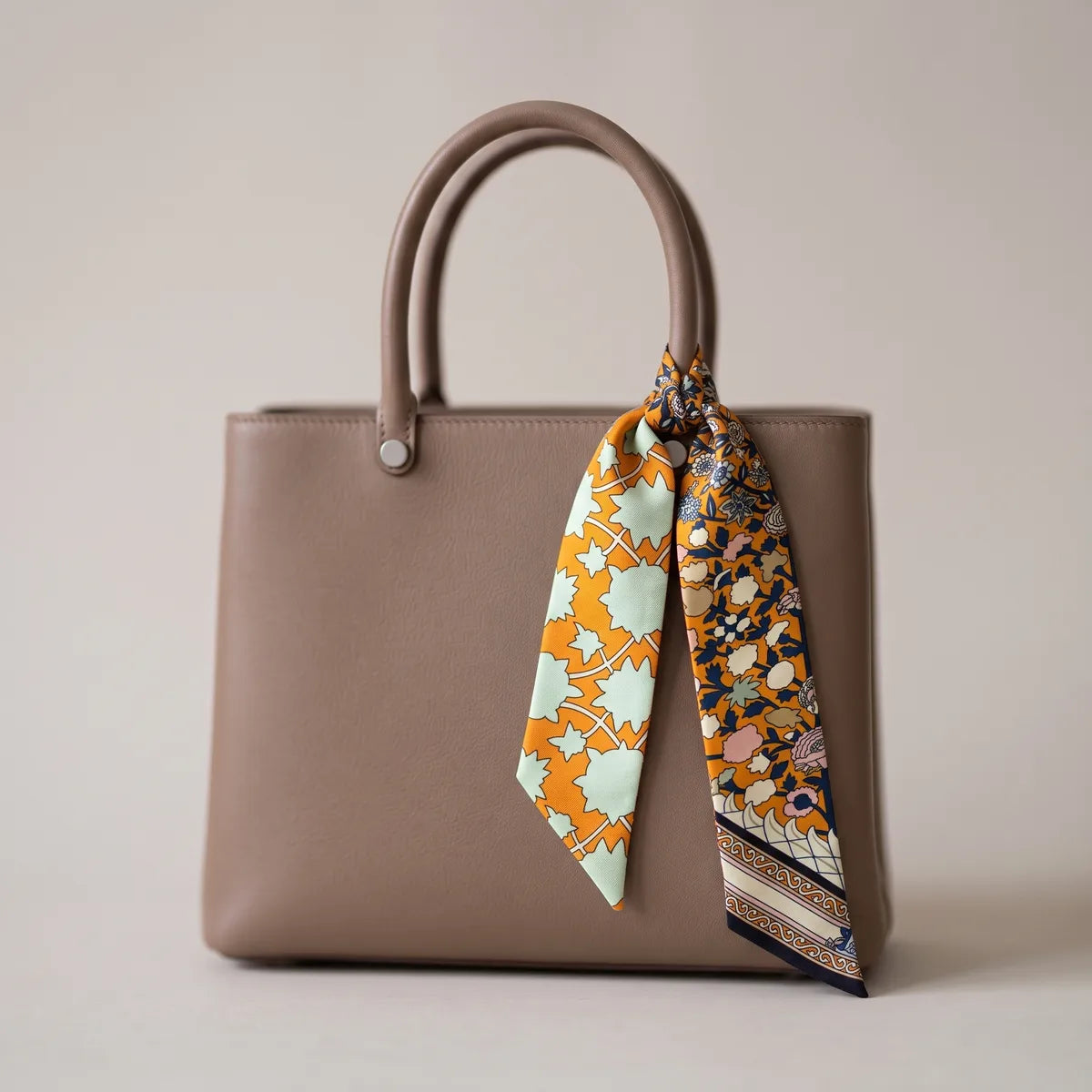 Orange and navy skinny silk scarf knotted around a taupe bag handle, tips hanging