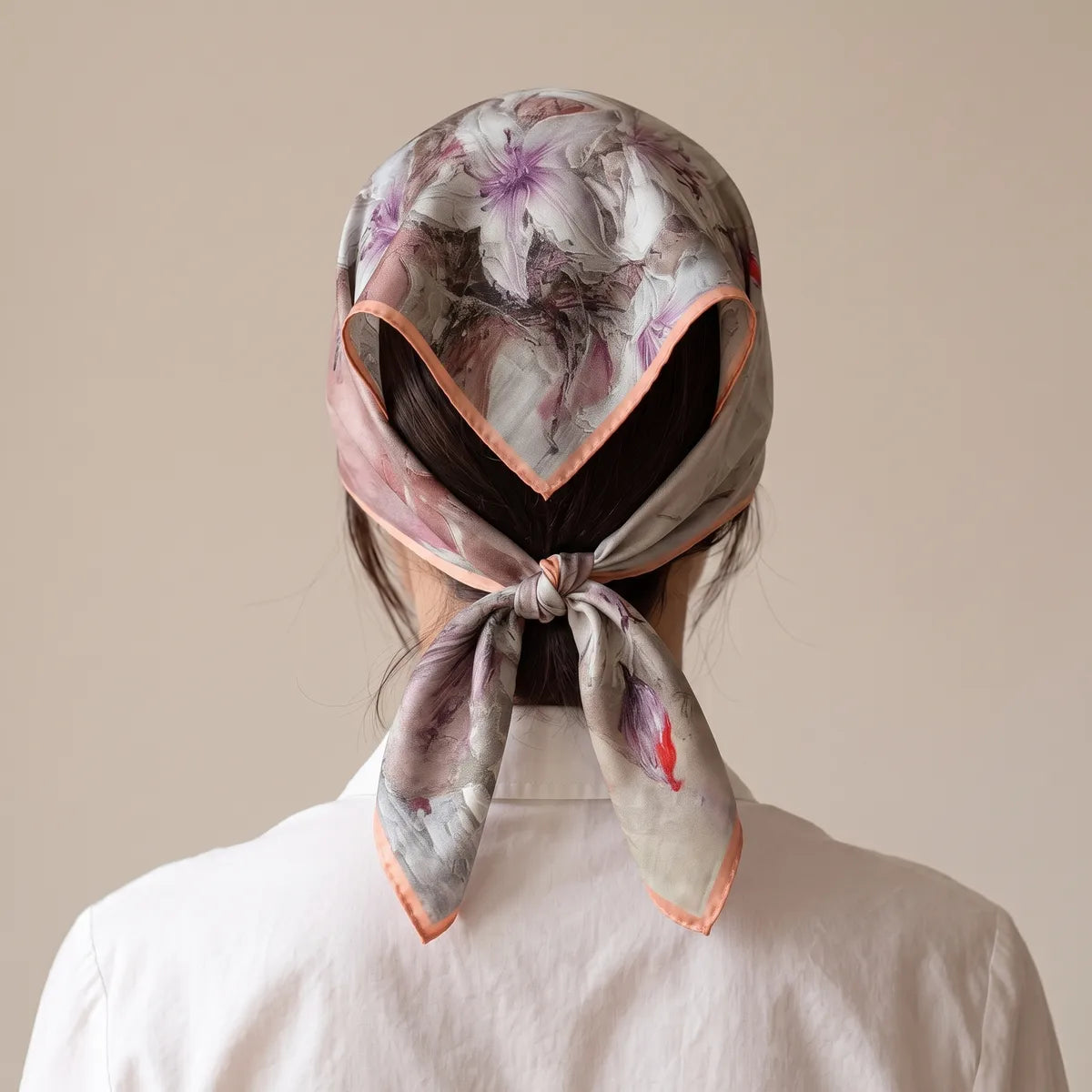 Back view of a pink lily silk scarf covering the hair, its coral-edged tails hanging from a knot at the nape
