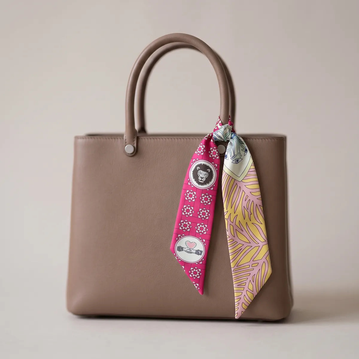 Pink medallion print skinny silk scarf knotted on a taupe bag handle, yellow leaves on one end