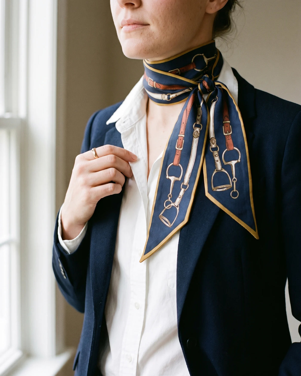Navy equestrian-print skinny silk scarf knotted at the neck over a white shirt and navy blazer