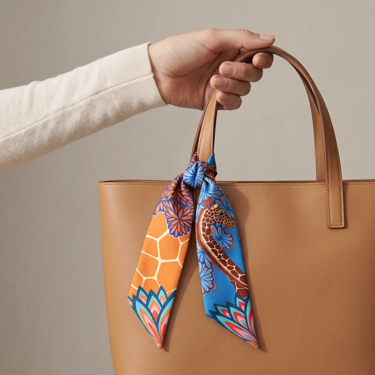 Blue giraffe print skinny silk scarf knotted on the handle of a tan leather tote