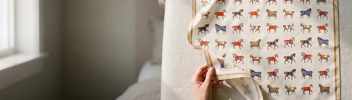 Hand lifting the corner of a cream horse print silk square laid on a linen bench