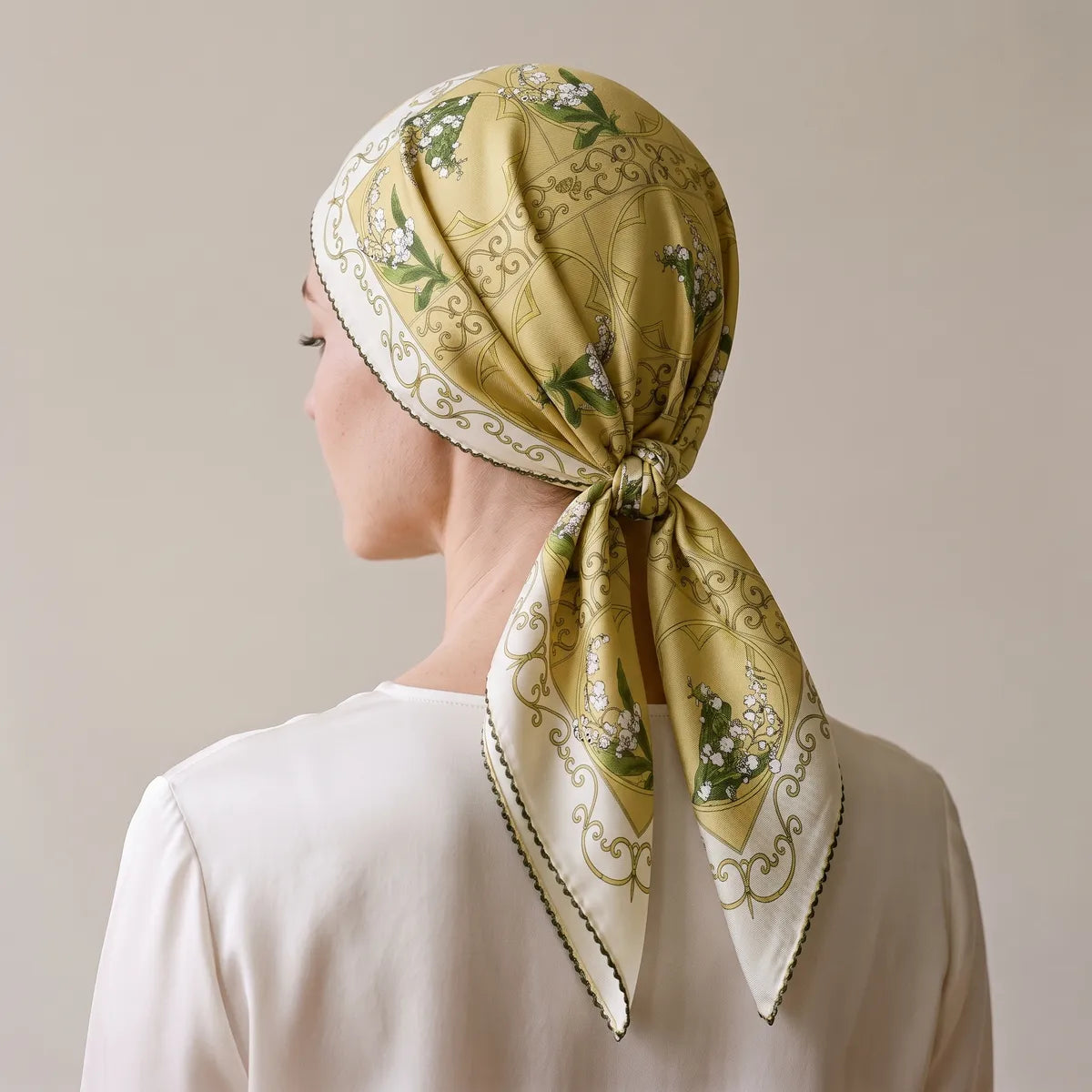 Yellow lily print scarf worn over the hair and knotted at the nape, seen from behind